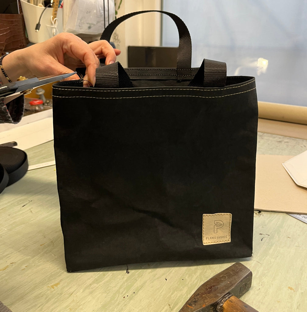 Hands of a craftswoman finishing a vegan leather lunch bag in a workshop, with scissors and hammer on the table – authentic moment of slow design and mindful craftsmanship by Plant Inside