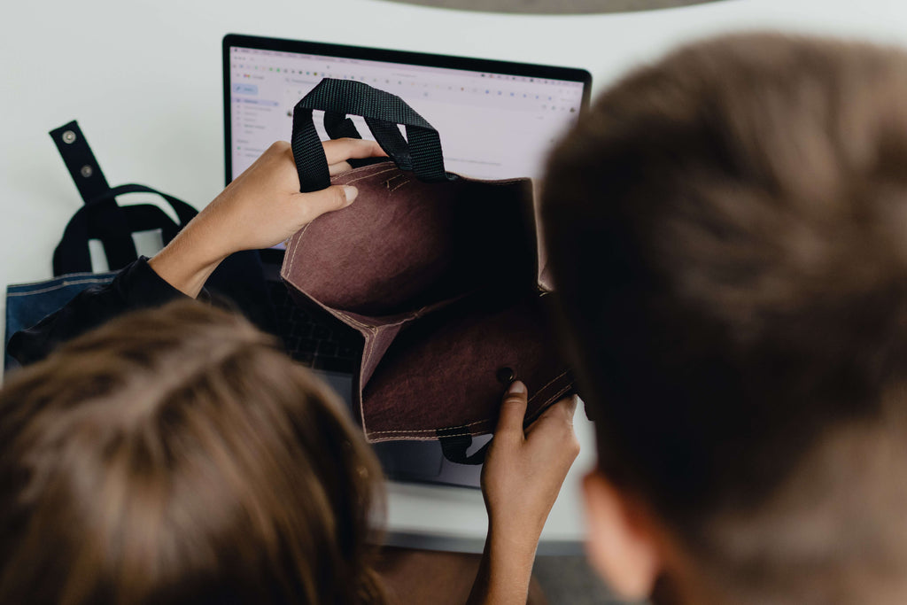 Top-down view of an open vegan lunch bag by Plant Inside in an office setting, with two colleagues discussing it and a laptop in the blurred background
