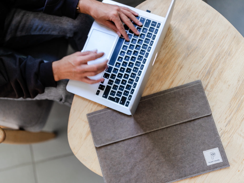 Top view of a minimalist office desk with a vegan laptop sleeve by Plant Inside, a MacBook, and hands typing – sustainable office fashion in action